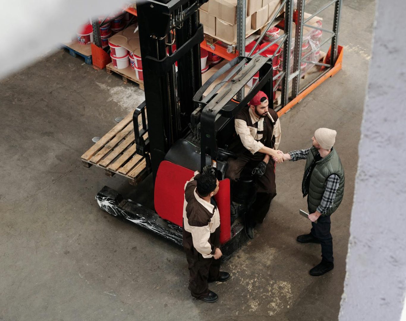 Three warehouse workers in conversation and handshake next to a forklift in an industrial setting.